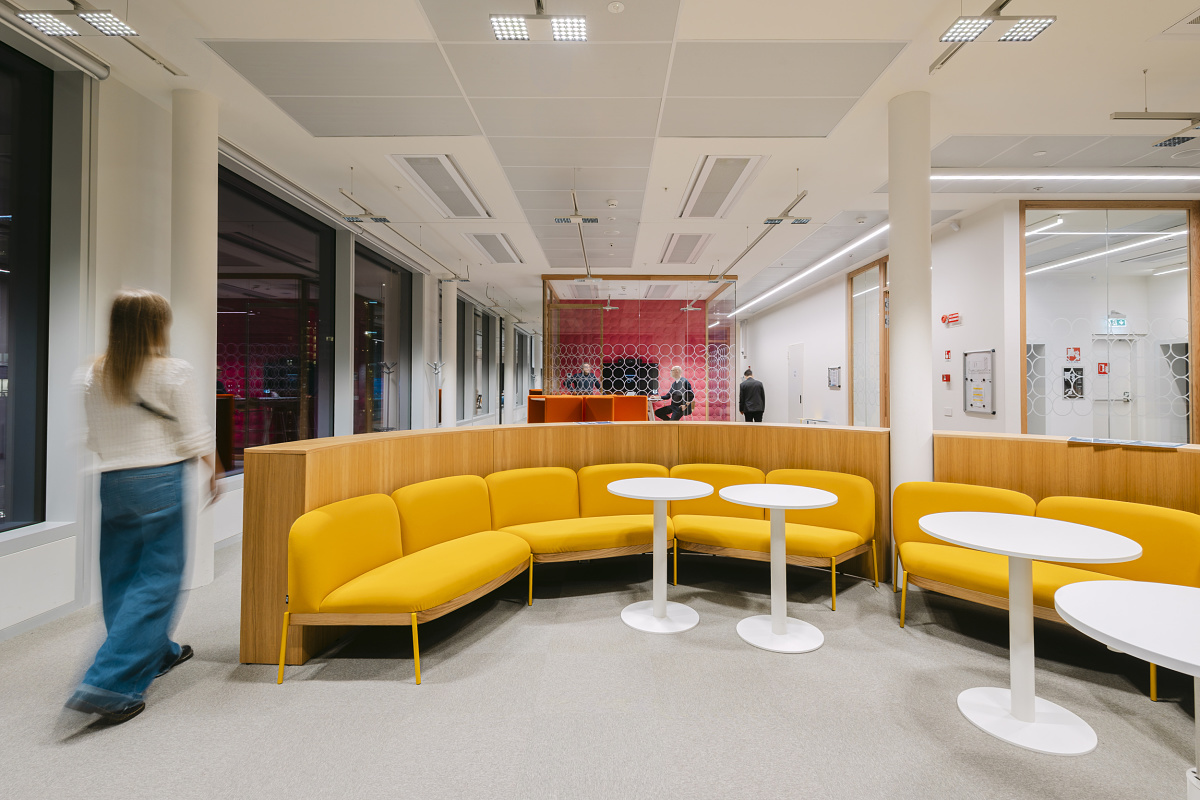 Modern office area with a curved yellow bench and white round tables in the foreground. In the background, a glass-walled meeting room with red wall panelling can be seen, with people moving through the room.