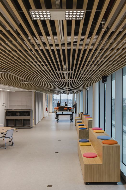 Large modern office space with a slatted timber ceiling, full-height windows and coloured seating steps. In the background are a table tennis table and high stools, where two people are seated by the window.