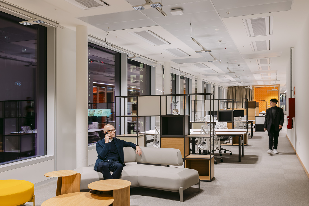 Modern office area with full-height windows, bright workstations, and a lounge sofa in the foreground. In the background, several desks with monitors are visible while a person walks through the room.