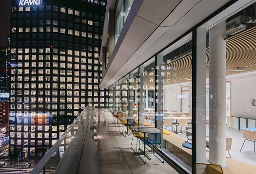 Night-time view of a modern office terrace with a glass balustrade and coloured chairs along the glazed facade. On the right, a bright interior seating area is visible; on the left stands a brightly lit office tower with numerous windows.