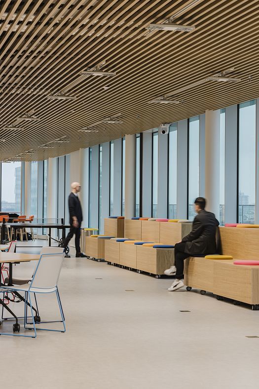 Large modern office space with floor-to-ceiling windows, a slatted timber ceiling and coloured seating steps. One person is seated on the steps, while another moves through the room.