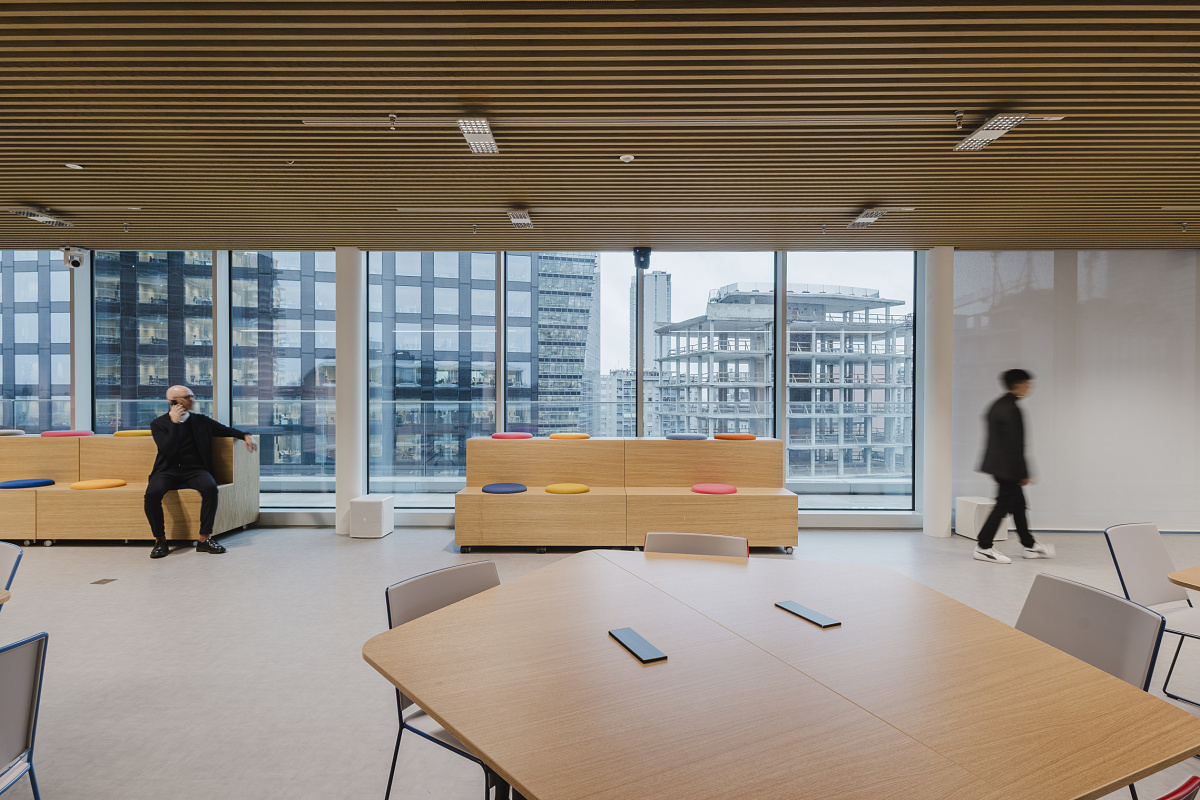 Bright office interior with large windows overlooking surrounding high-rise buildings. In the foreground, timber tables and chairs are grouped together; in the background, one person sits on coloured seating steps while another walks through the room.