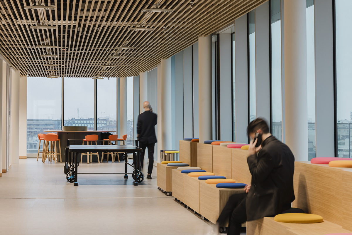 Bright office interior with large windows, a slatted timber ceiling, coloured seating steps and a table tennis table. One person is seated on the steps talking on the phone, while another walks through the room.