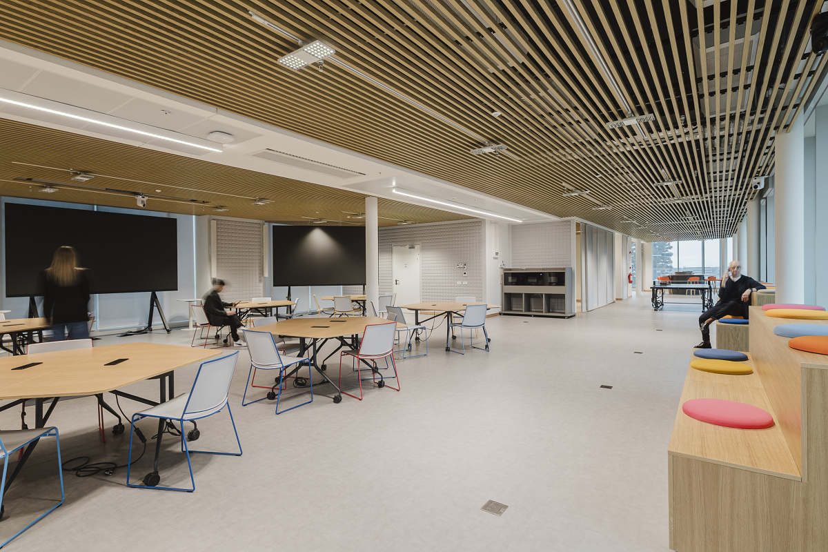 Large open-plan office with a slatted timber ceiling, several round desks and large monitors. On the right, coloured seating steps sit beside full-height windows, with one person seated there while others work or move through the room.
