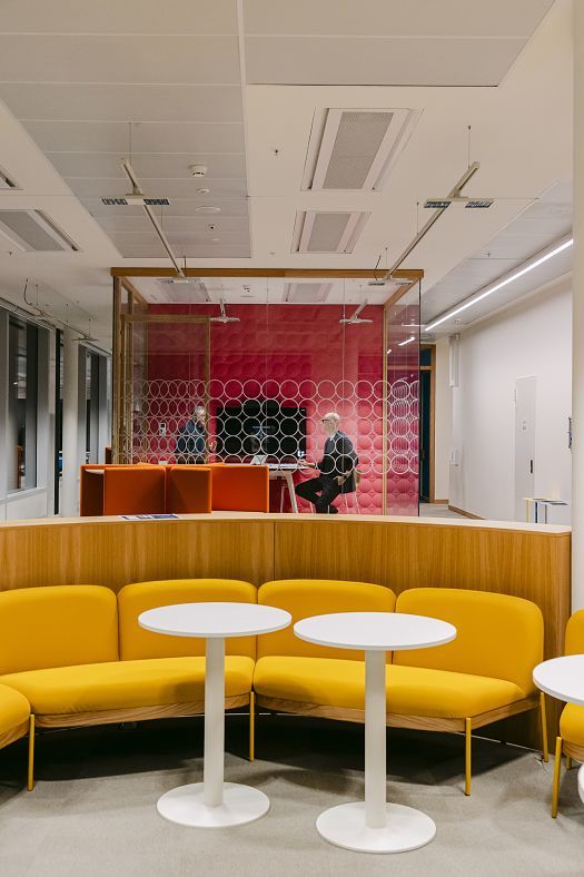 Modern office area with yellow benches and white round tables in the foreground. Behind them is a glass-walled meeting room with red wall panelling and two people seated at a table.
