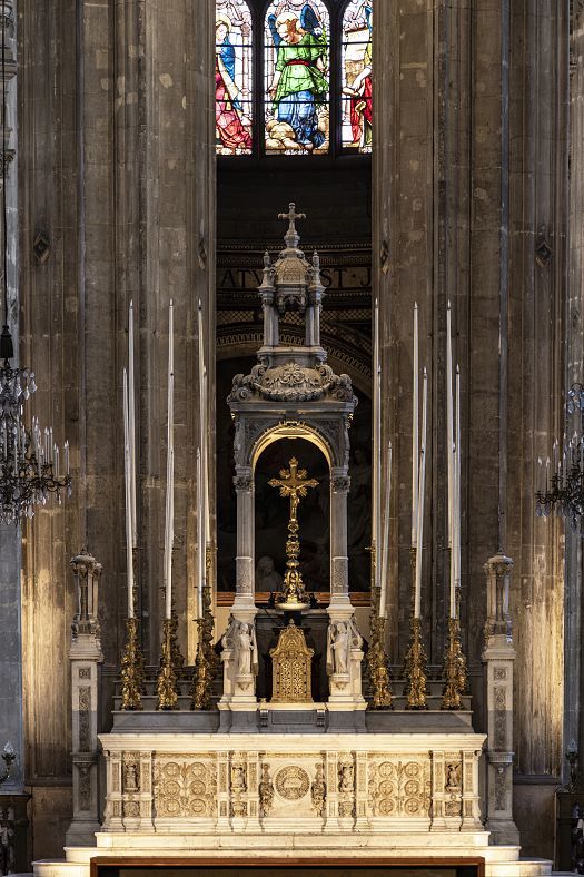 Close-up view of an ornate church altar made of light-colored stone, featuring tall candlesticks, intricate carvings, and a central tabernacle topped with sculptural details. Above the altar, a stained-glass window with colorful figures is visible, framed by tall stone columns and chandeliers on either side.