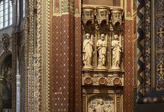 Detailed view of an ornate sculptural wall inside a historic church, featuring carved stone figures standing in recessed niches beneath a richly decorated canopy. The surrounding wall is covered with intricate geometric patterns in red, gold, and green, with additional relief carvings visible below the statues.