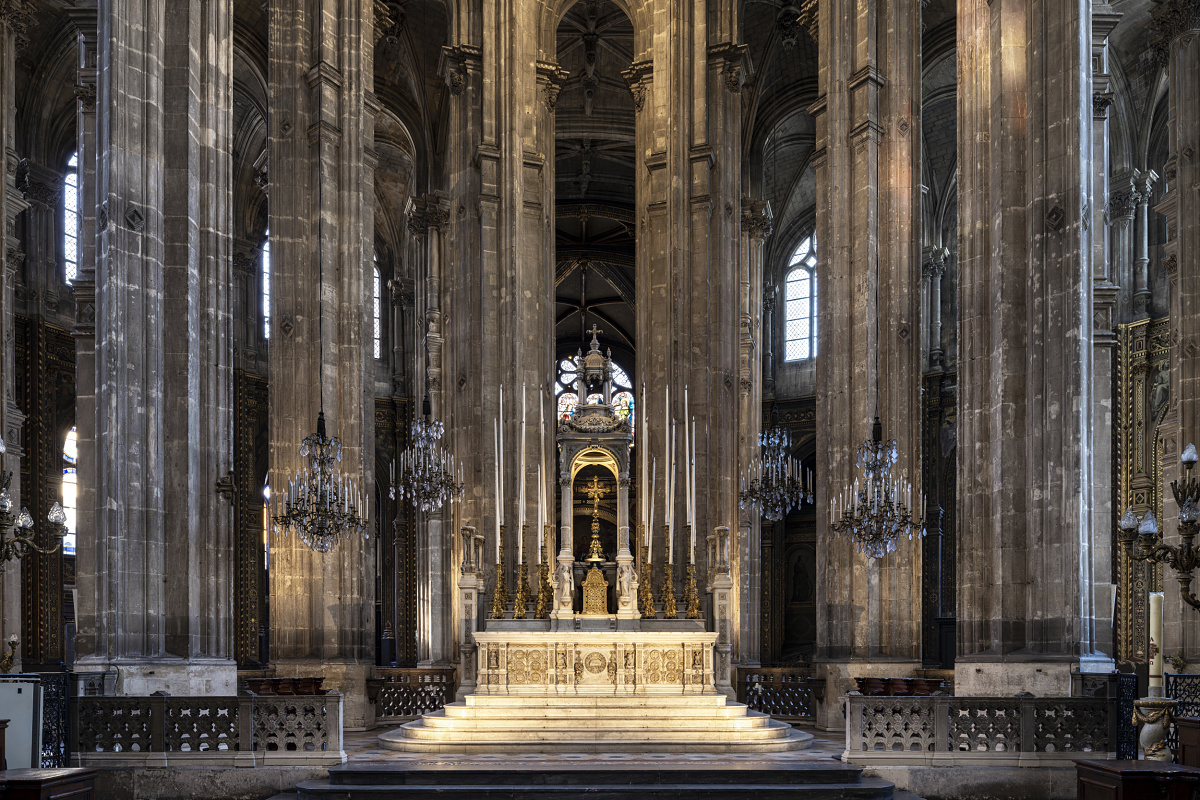 Front view of a grand church altar framed by tall stone columns, ornate chandeliers, and intricate Gothic architectural details. The illuminated altar, with carved decorations and tall candlesticks, stands at the center beneath high vaulted arches and large arched windows.